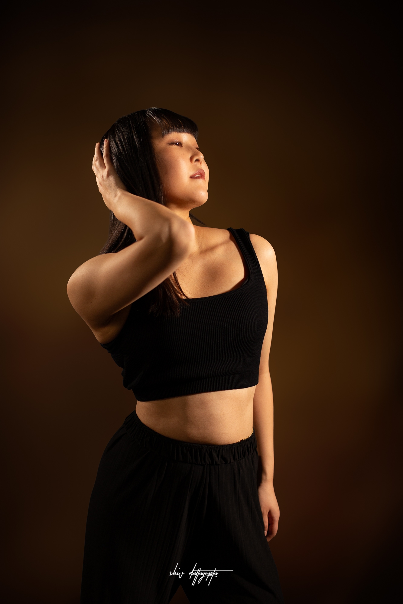 Portrait of a woman with long black hair in a black crop top, her hand in her hair against a warm brown studio backdrop.